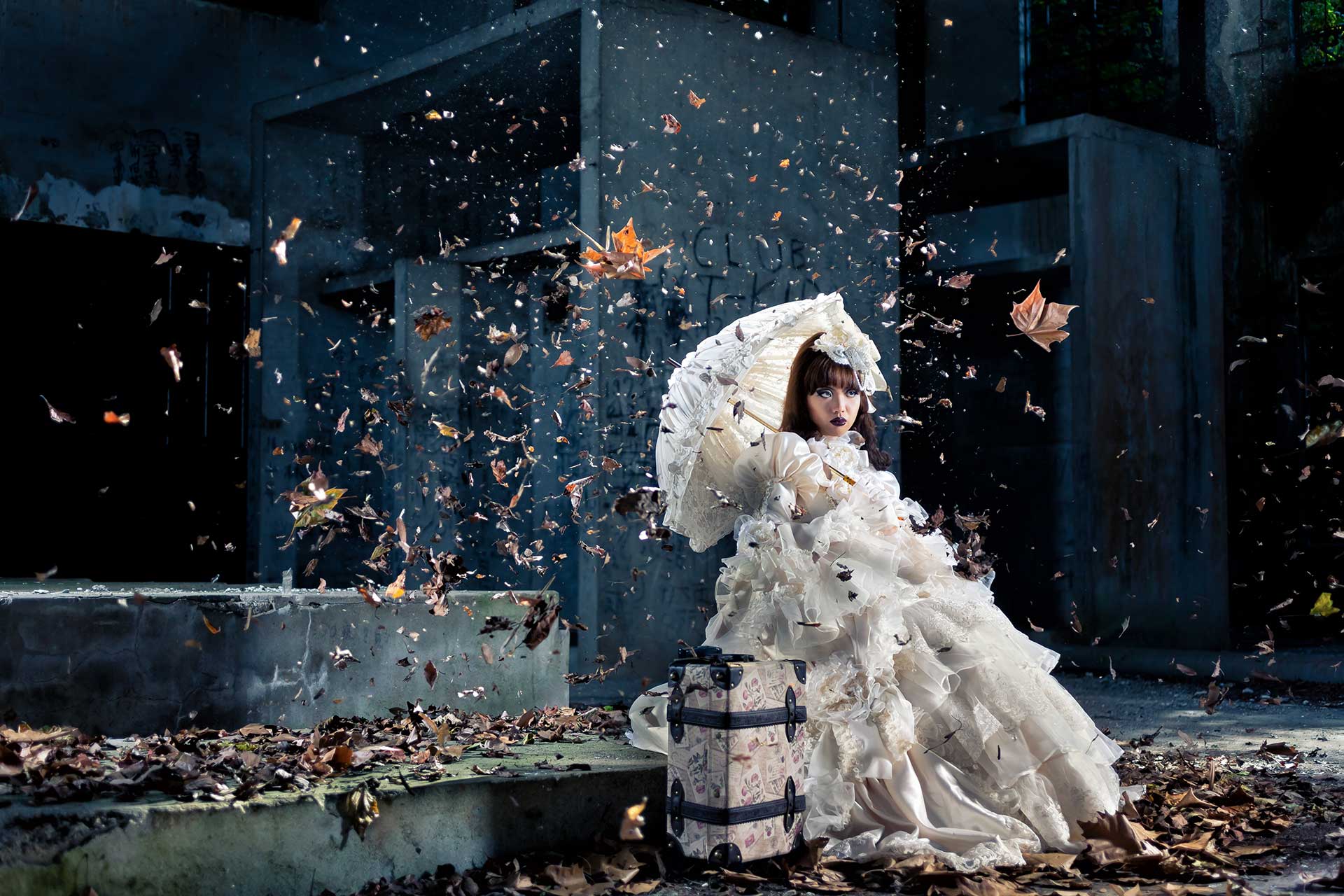 Bride in white dress sitting on ground with falling leaves, part of groom creative pre-wedding photoshoot.